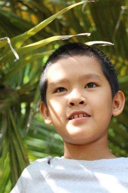 Neşeli genç bir çocuk yemyeşil yaprakların altında durarak sıcak güneş ışığında masumiyet ve mutluluk saçarak güvenle gülümsüyor. Doğa odaklı temalar için mükemmel.