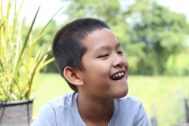 Gülümseyen neşeli bir genç çocuk açık havada güneşli bir günün tadını çıkarıyor. Yeşillik ve yemyeşil bir zemine dayanan bu samimi portre, çocukluk mutluluğunun ve oyunculuğunun özünü yakalar..