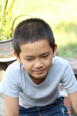 Düşünceli genç bir çocuk dışarıda oturuyor, doğal ışık ve yeşillikle çevrili. Onun gevşek ifadesi, çocukluğun masumiyetini huzurlu bir anda yakalar..