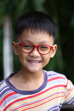 Gözlüklü neşeli bir çocuk renkli çizgili bir gömlek giyerken geniş bir şekilde gülümsüyor. Görüntü onun neşesini ve neşesini doğal bir şekilde yansıtıyor..