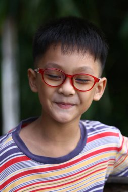 Kırmızı gözlüklü ve çizgili tişörtlü neşeli bir çocuk sıcak bir şekilde gülümsüyor. Görüntü, masumiyet ve mutluluğu doğal bir ortamda yakalar..