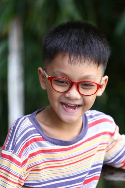 Kırmızı gözlüklü ve renkli çizgili tişörtlü neşeli bir çocuk açık hava ortamında parlak bir şekilde gülümsüyor, saf mutluluk ve masumiyet anını yakalıyor..