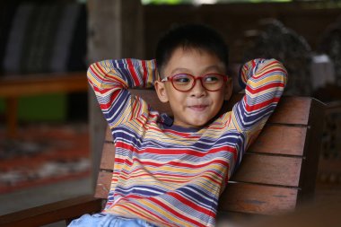 Rahat genç bir çocuk ahşap bir sandalyede rahat bir şekilde oturuyor, renkli çizgili bir kazak ve gözlük takıyor, neşe ve neşe saçıyor..