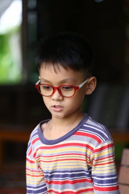 Kırmızı gözlüklü, renkli çizgili tişörtlü, kapalı bir yerde yakalanmış düşünceli genç bir çocuk. Odaklanmış ifadesi merak ve derinliği yansıtıyor..