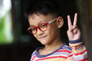 Şık gözlük takan neşeli bir çocuk barış işaretiyle poz veriyor, neşe ve masumiyet yayıyor. Çocukluk ve mutluluk temaları için mükemmel..
