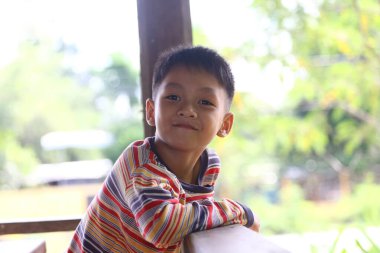 Çekici genç bir çocuk dışarıda otururken parlak bir şekilde gülümsüyor. Doğal ışık onun neşeli yüz ifadesini güçlendirir, çocukluk sevincinin ve masumiyetinin özünü yakalar..