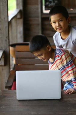 İki çocuk, ahşap bir masada, rahat bir ortamda dizüstü bilgisayarla vakit geçirmenin keyfini çıkarıyorlar..