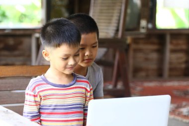 İki çocuk bir dizüstü bilgisayarın etrafında toplanıyor, evlerinde eğitici aktiviteler yapıyorlar, sıcak ve davetkar bir atmosferde merak ve takım çalışması sergiliyorlar..