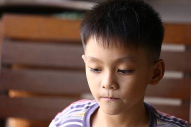 Kısa saçlı düşünceli genç bir çocuk evde oturur, ciddi bir ifadeyle bakar. Çizgili gömleği sakin bir ana renk katıyor..