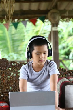 Yoğunlaşmış genç bir çocuk kulaklığı takmış, yeşil ve kültürel dekorlarla çevrili rahat bir iç mekan ortamında dizüstü bilgisayarıyla meşgul..