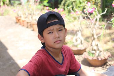 Dışarıda günlük kıyafetlerle oturan genç bir çocuk, düşünceli bir yüz ifadesi sergiliyor. Yeşilliklerle çevrili bu samimi anda masumiyet ve merak yayıyor..