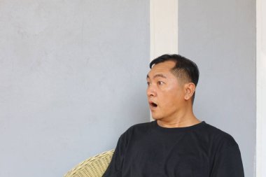 Şaşırmış bir adam evde otururken şok olmuş bir yüz ifadesi sergiler. Günlük kıyafetleri ve rahat duruşu o anın gerçekliğine katkı sağlıyor, kişisel duygularını gösteriyor..