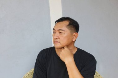 Düşünceli bir adam rahat bir ortamda oturur, çenesini eline koyar, gri bir duvara karşı düşünceli bir yüz ifadesi sergiler. Portre, iç gözlem ve sakinliği yansıtıyor..