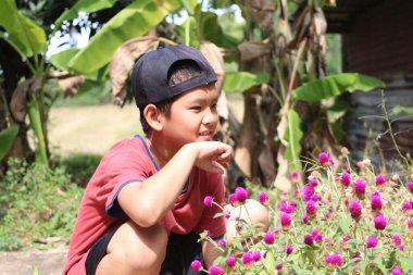 Genç bir çocuk renkli çiçeklerin arasında oturmuş, sakin bir doğal ortamda merak ve neşe sergiliyor, çocukluk keşfi ve mutluluğunu yakalıyor..