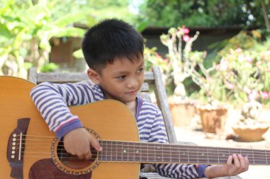 Genç bir çocuk sakin bir açık hava ortamında akustik gitar çalmayı seviyor. Müzikal yeteneğini ve neşeli ifadesini doğal bir arka plana karşı sergiliyor..