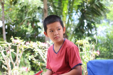 Genç bir çocuk, yemyeşil ve renkli çiçeklerle dolu canlı bir bahçede sakince oturur, çocukluk masumiyetinin ve keşiflerinin özünü yakalar..