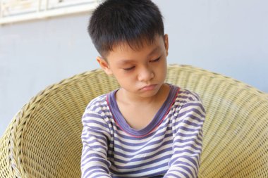 Çizgili gömlekli genç bir çocuk örülmüş bir sandalyede düşünceli bir şekilde oturur. Düşünceli ifadesi, kapalı alanda sessiz bir yansıma ve masumiyet anı yakalıyor..