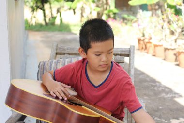 Dışarıda oturan genç bir çocuk gitar çalmakla meşgul. Yeşilliklerle çevrelenmiş halde müzik yapma deneyiminin tadını çıkarırken odak ve yaratıcılık sergiliyor..