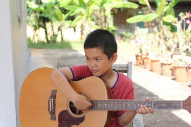 Dışarıda oturan genç bir çocuk, akustik gitarını çalmaya odaklanmış. Sahne, huzurlu bir bahçe ortamında müzik ve yaratıcılık dolu bir anı yakalıyor..