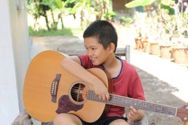Neşeli bir çocuk açık havada oturuyor, parlak bir gülümsemeyle akustik gitarını çalıyor. Sahne çocukluk sevincinin ve müzik yeteneğinin özünü doğal bir ortamda yakalıyor..