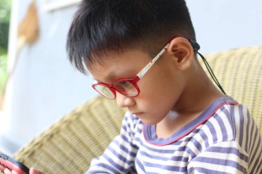 Kırmızı gözlüklü genç bir çocuk akıllı telefonuna odaklanmış, modern çocukluk anılarını gözler önüne seriyor..