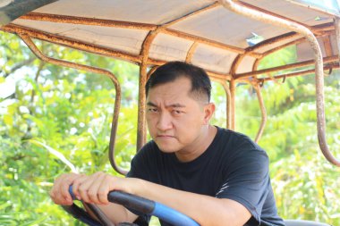 Bir adam yemyeşil bir ortamda traktör kullanırken ciddi bir yüz ifadesi sergiler. Görüntü onun odaklanmasını ve doğadaki kararlılığını yakalıyor..