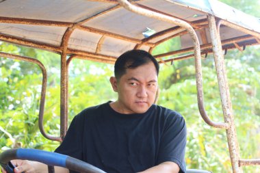 Traktör kullanan bir adam kararlılığını odaklanmış bir ifadeyle gösterir. Yeşillik ile çevrelenmiş, kırsal tarım ruhunu somutlaştırıyor..