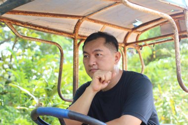 Düşünceli bir adam canlı yeşilliklerle çevrili taşralı bir araçta oturuyor. Dalgın görünüyor, doğadaki huzurlu anın tadını çıkarıyor..