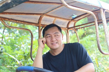 Neşeli bir adam, yemyeşil bir ortam içindeyken, bir araçta oturup, çenesini avucunun üzerinde dinlendirirken, doğadaki rahatlama ve neşeyi yansıtıyor..