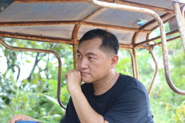 Düşünceli bir adam paslı bir yapının altında oturuyor, yeşil, doğal bir ortamda dalgın dalgın dalgın yatıyor. Yüzündeki ifade derin duyguları ve rahatlığı yansıtıyor..