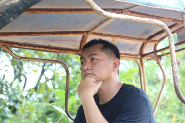 Düşünceli bir adam kırsal bir yerde, yemyeşil ortamda dalgın dalgın dalgın oturuyor. Düşünceli ifadesi iç gözlem ve huzur dolu bir anı yansıtıyor..