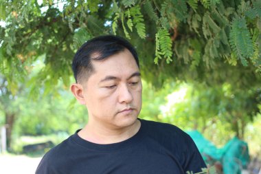 Bu görüntüde siyah gömlekli bir adam yeşil ağaçların altında düşünceli bir şekilde yansıyor, aydınlık bir günde doğadaki huzurlu bir anı yakalıyor..