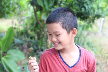 Neşeli bir çocuk küçük yeşil bir bitkiyi tutarken gülümsüyor, doğanın sıcaklığının tadını çıkarıyor. Bu samimi an, masumiyeti ve merakı yemyeşil bir ortamda yakalar..