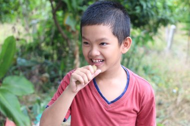 Neşeli bir çocuk dışarıda eğleniyor, küçük bir atıştırmalık tutarken gülümsüyor. Çevresindeki canlı doğa, çocukluğun neşeli atmosferine katkı sağlıyor..