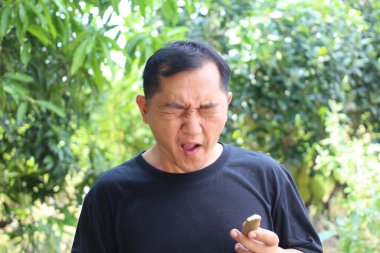 Açık havada bir adam meyveyi tutarken tiksindirici bir ifade sergiler. Yeşilliklerle çevrelenmiş bu görüntü, gerçek duyguları ve doğayla eğlenceli etkileşimleri yakalar..