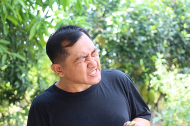 Açık hava ortamında bir adam acı dolu bir yüz ifadesi sergiliyor. Yeşilliklerle çevrili, rahatsızlık ve hüsranla dolu duygular taşır..