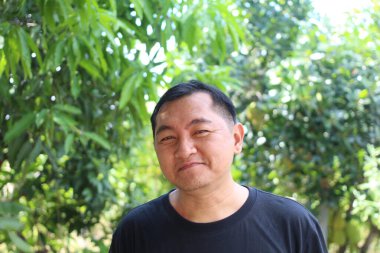 Neşeli bir adam, yemyeşil yeşilliklerle çevrili canlı bir ortamda sıcak bir şekilde gülümsüyor. Sahne, doğada bir mutluluk ve sükunet anı yakalar..