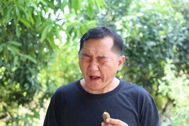 Bir adam yemyeşil bir bahçede meyve tadarken dramatik bir tepki verir. Yüzündeki ifade şaşkınlık ve hoşnutsuzluk anını yakalıyor. Taze yemek tatma sevincini gözler önüne seriyor..