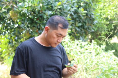 Bir adam canlı bir bahçede yeşil bir yaprağı yakından inceliyor. Sahne, doğayla olan bağlantısını ve bereketli bir ortamda düşünme anını yansıtıyor..