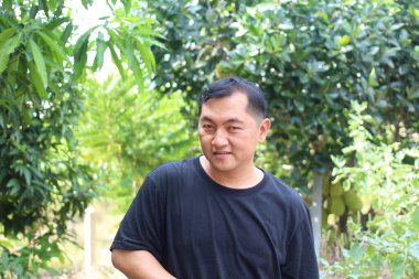 Siyah gömlekli bir adam yemyeşil bir ortamda poz veriyor, etrafındaki barışçıl doğanın tadını çıkarıyor. Bu canlı ortam açık havada neşeli bir anı yakalıyor..