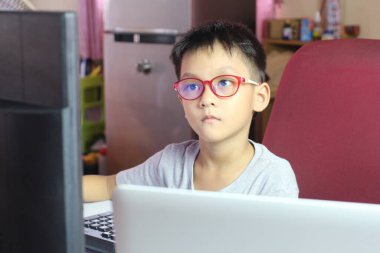 Gözlük takan odaklanmış bir çocuk bilgisayarın başında oturur ve öğrenmeye derinden bağlıdır. Bu görüntü, gençliğin ve teknolojinin özünü bir ev çalışma ortamında yakalıyor..