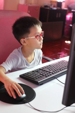 Gözlüklü odaklanmış bir çocuk bilgisayar masasında oturuyor, ekranıyla meşgul. Parlak iç mekan, modern öğrenme ve keşiflerin özünü yakalar..