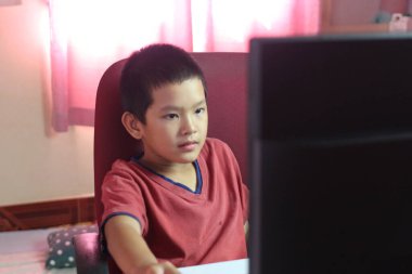 Genç bir çocuk bilgisayar ekranının önünde oturur, elindeki işe derinden odaklanır. İçerideki rahat ortam yumuşak ışıklandırma ve sıradan bir atmosfer içeriyor..