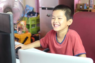 Neşeli bir çocuk bir masada oturuyor, mutlu bir şekilde bilgisayarla meşgul. Gülüşü neşe ve merakı yansıtıyor. Çocukluğunun enerjisini bir yuva ortamında sergiliyor..