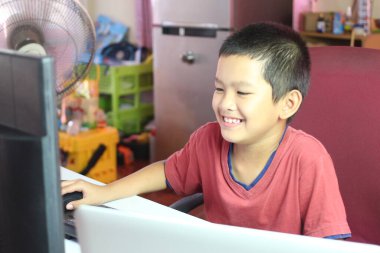 Neşeli bir çocuk evde bilgisayar kullanırken gülümsüyor. Görüntü, çocukların dijital çağdaki sevinç ve merakını yakalıyor. Teknolojinin öğrenmedeki rolünü gösteriyor..