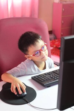 Gözlüklü genç bir çocuk masada oturur, bilgisayar kullanmaya odaklanır. Parlak oda ve neşeli atmosfer yaratıcılığa ve öğrenmeye ilham verir..