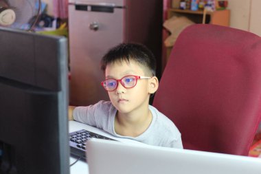 Gözlük takan genç bir çocuk bilgisayar ekranına odaklanmış bir şekilde bir masada oturuyor. Kapalı alan, modern bir ev ortamını yansıtıyor..