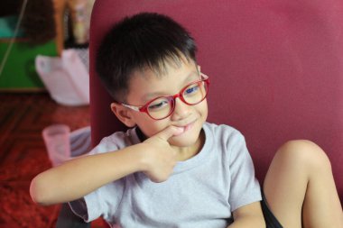 Kırmızı gözlüklü neşeli bir çocuk rahatça oturur, eliyle şakacı hareketler yaparken düşünür. İçerideki sıcak ortam, sıcak atmosfere katkı sağlıyor..