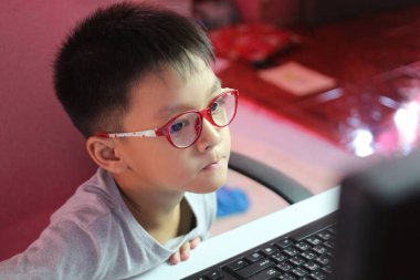 Gözlüklü bir çocuk dikkatle bilgisayar ekranına odaklanıyor ve evdeki modern öğrenme ortamını gösteriyor. Eğitim temaları için mükemmel.