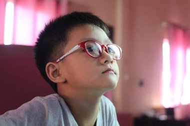 Kırmızı gözlüklü genç bir çocuk dikkatle bakıyor, düşüncelere dalmış. Parlak aydınlatma, dingin atmosferi yükseltir, merakı ve dikkati gösterir..